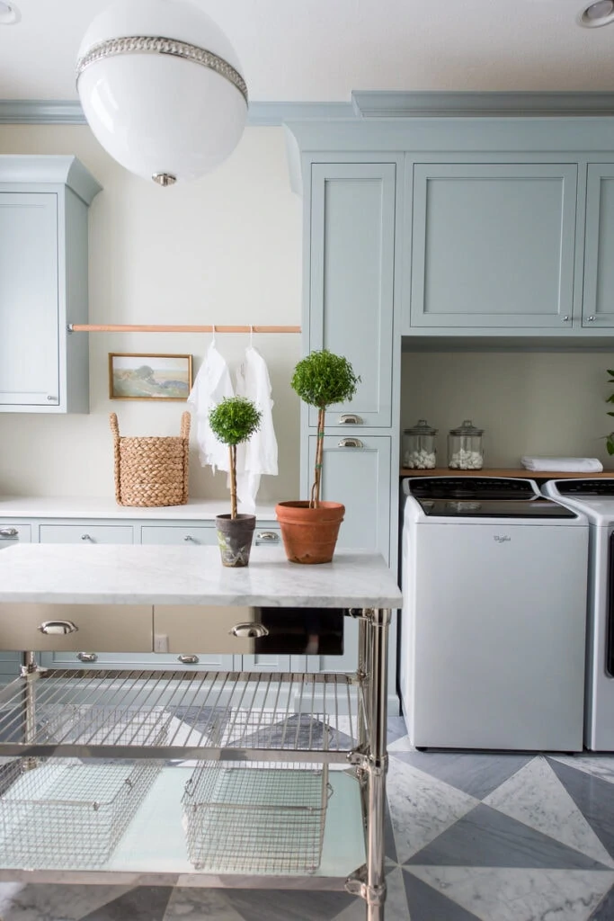 A gorgeous laundry room renovation featuring checkered marble floors, light blue cabinets, a round light fixture, and topiaries on a silver cabinet base with marble top!