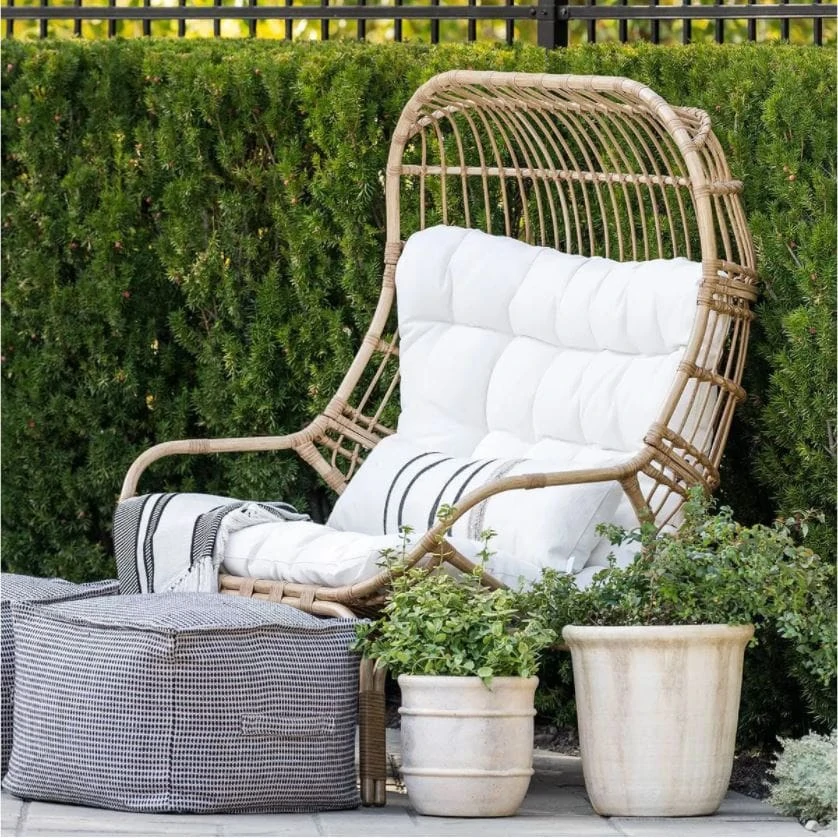 An outdoor egg chair with lumbar pillow, outdoor blanket, poufs and concrete planters from the new Outdoor dining with striped outdoor wicker chairs, an outdoor wood dining table, and outdoor place settings from the Studio McGee Threshold Patio Collection.