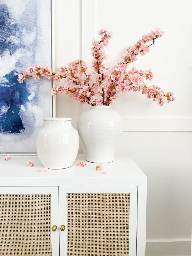 Spring decorating ideas in a home office including faux cherry blossom stems in a large white urn on a white enamel and cane cabinet.