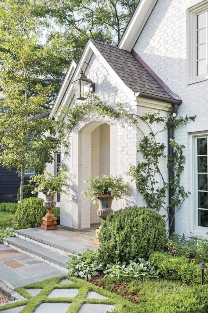 A white painted brick house exterior with brown roof, green climbing plants and white window trim.