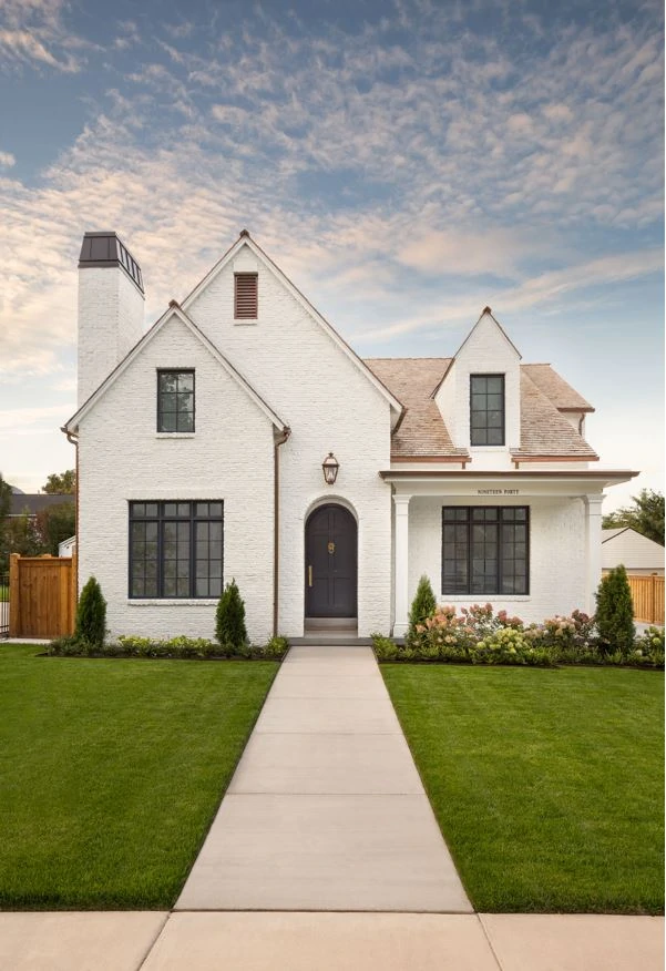 A Tudor exterior with white painted brick, black window trim, copper downspouts and a black arched front door.