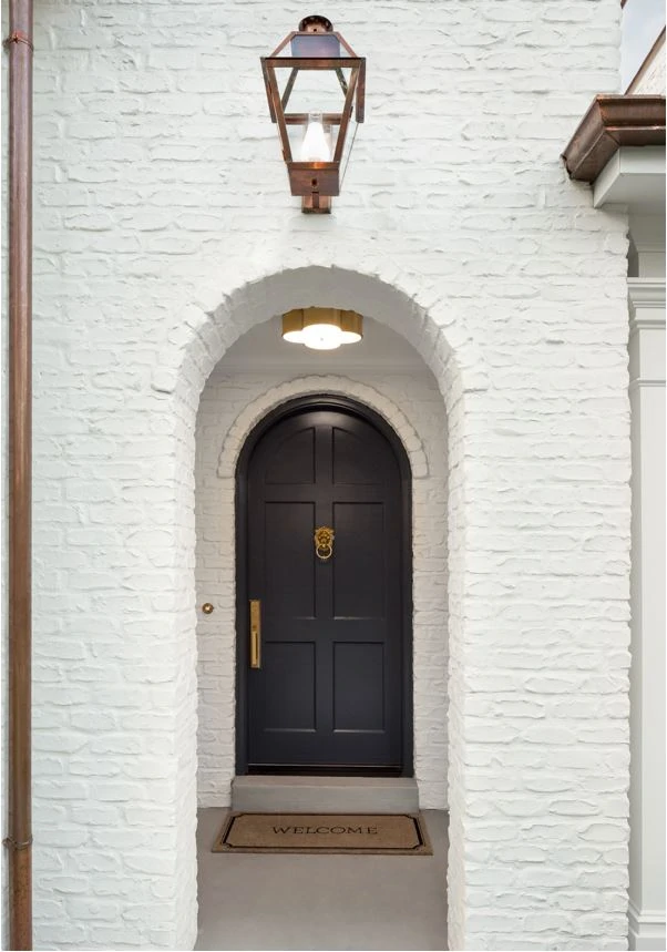 A white painted brick Tudor house with arched doorways, black front door, and copper downspouts.