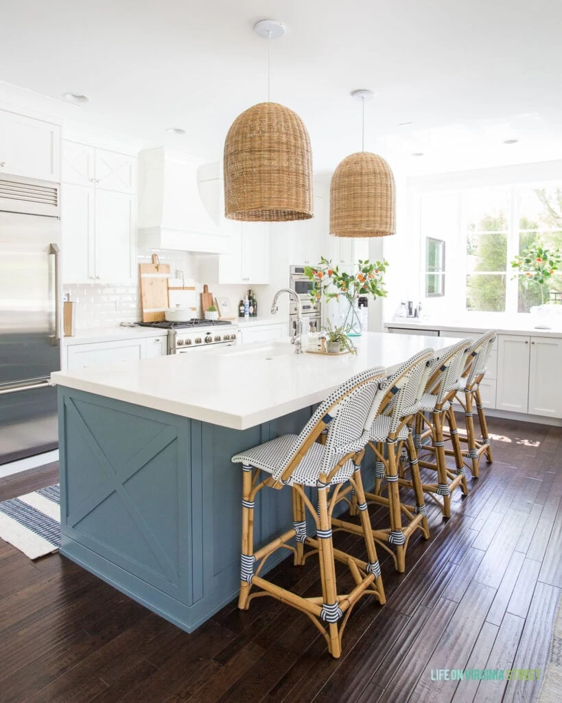 A kitchen with Benjamin Moore Simply White cabinets, Providence Blue island, woven basket pendant lights, bistro counter stools, a striped rug and faux orange stems.