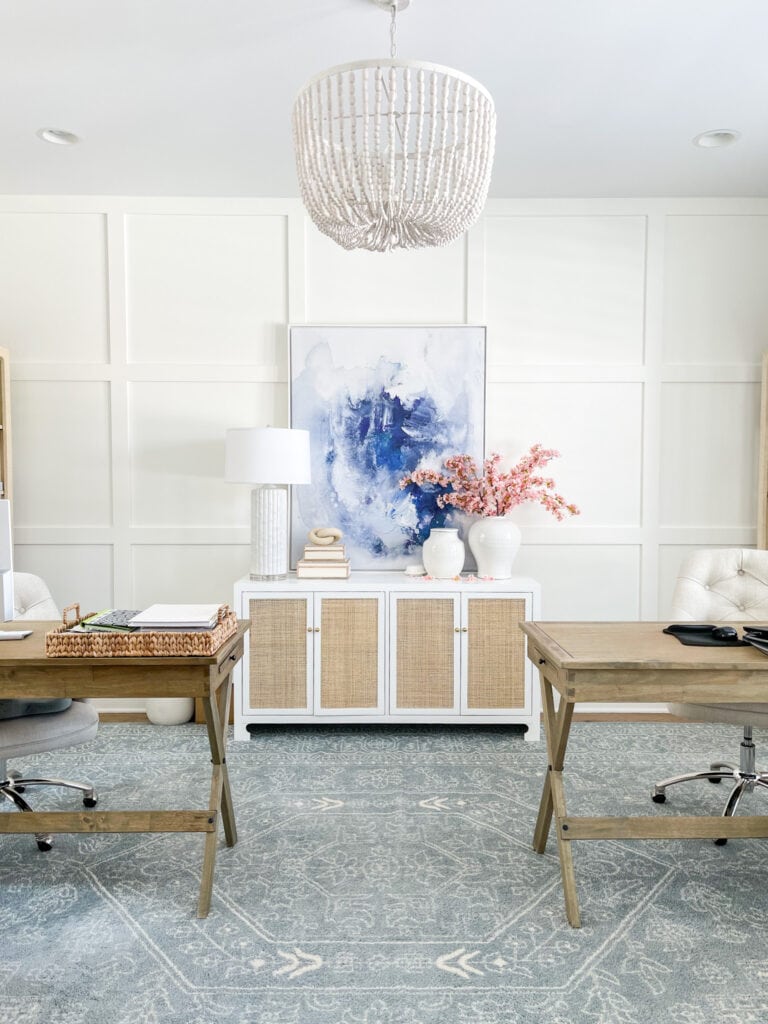 Home office with board and batten grid walls, wood campaign desks, a blue patterned rug, cane cabinet and large abstract art.