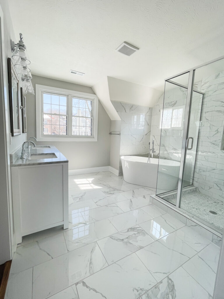 Master bathroom with affordable ceramic tile that looks like Carrara marble! Includes a soaking tub, glass vanity lights, multiple windows, large glass framed shower, and white double vanity.