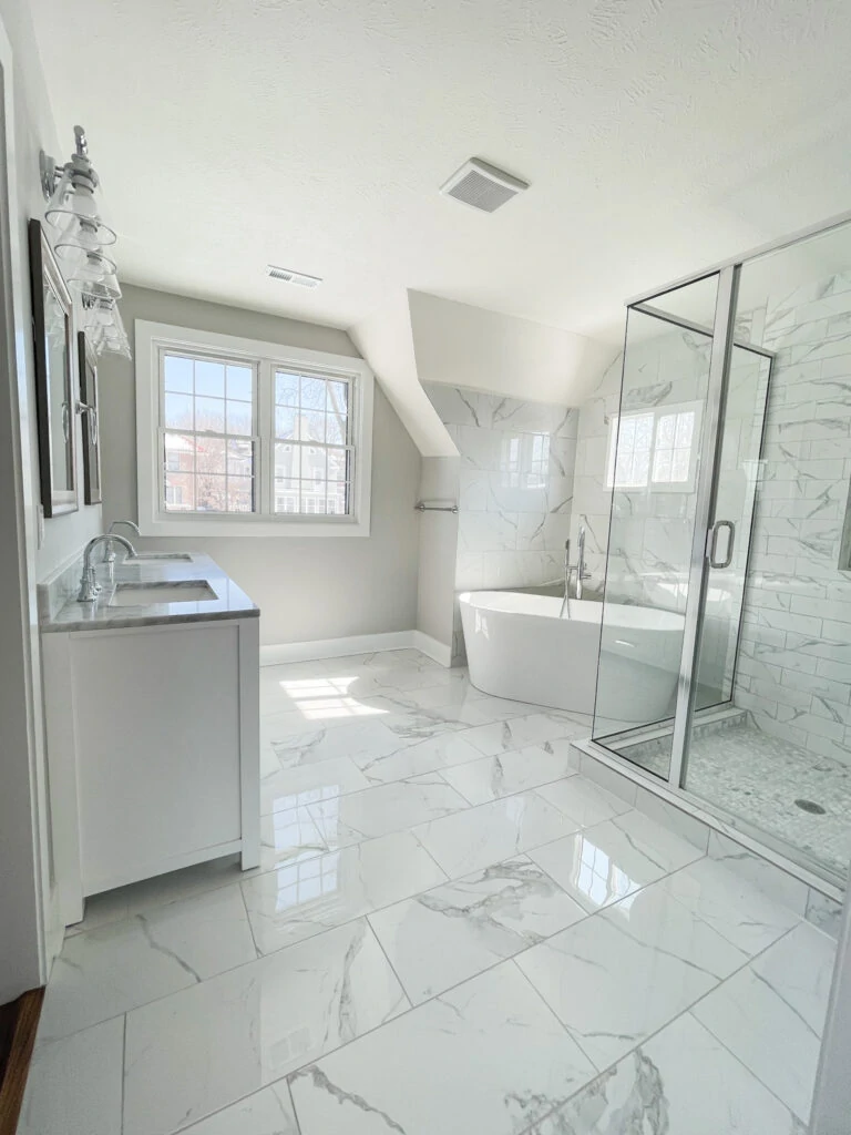 Master bathroom with affordable ceramic tile that looks like Carrara marble! Includes a soaking tub, glass vanity lights, multiple windows, large glass framed shower, and white double vanity.