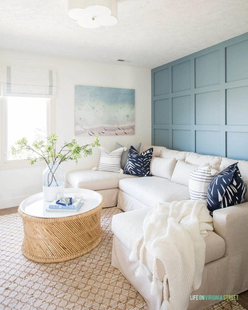 A TV room with blue board and batten grid wall with linen sectional, round coffee table and coastal wall art with a beach scene.
