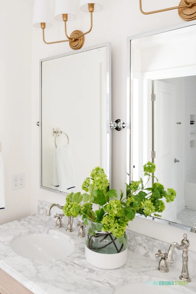 A Jack & Jill bathroom remodel reveal with Carrara marble countertops, polished nickel pivot mirrors, gold light fixtures, polished nickel faucets and faux viburnum stems.