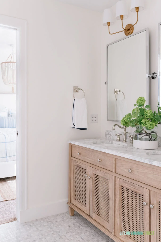 A Jack and Jill bathroom remodel that used wood cane vanity, Carrara marble floors, silver pivot mirrors, gold light fixtures and blue and green accents. You can see the bedroom in the background.