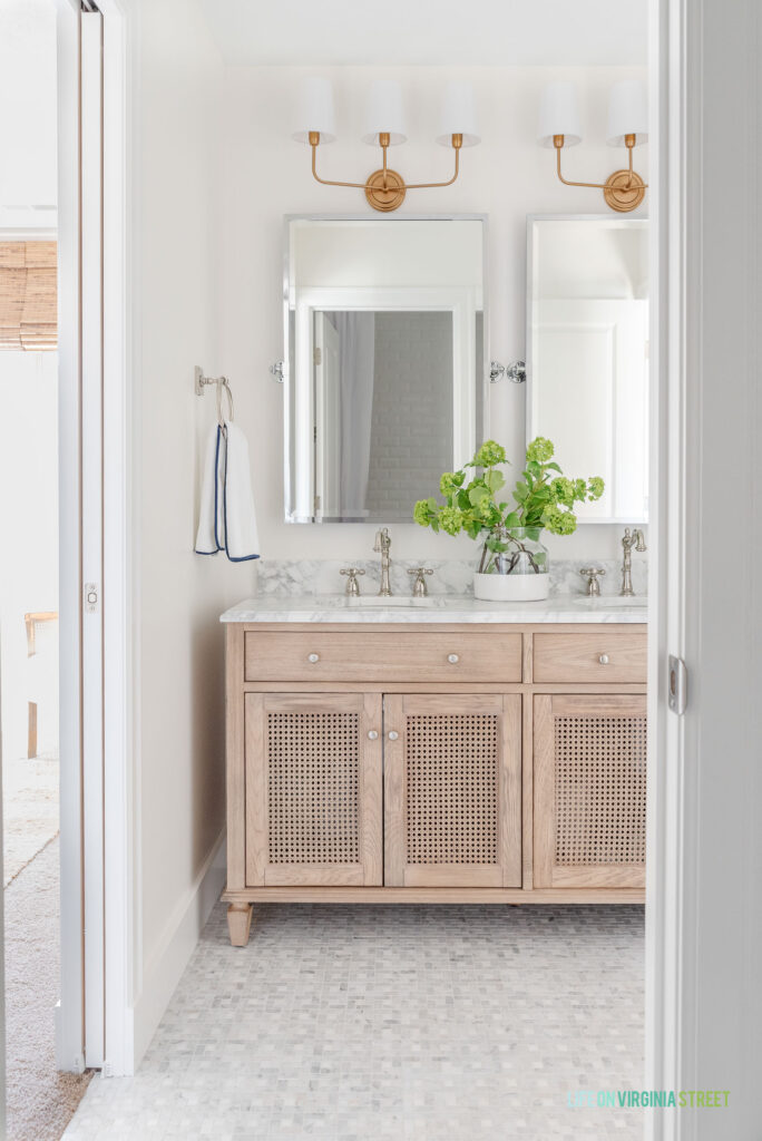 Gold bathroom vanity light fixtures hang above polished nickel pivor mirrors and a wood cane vanity. The floor is Carrara marble.