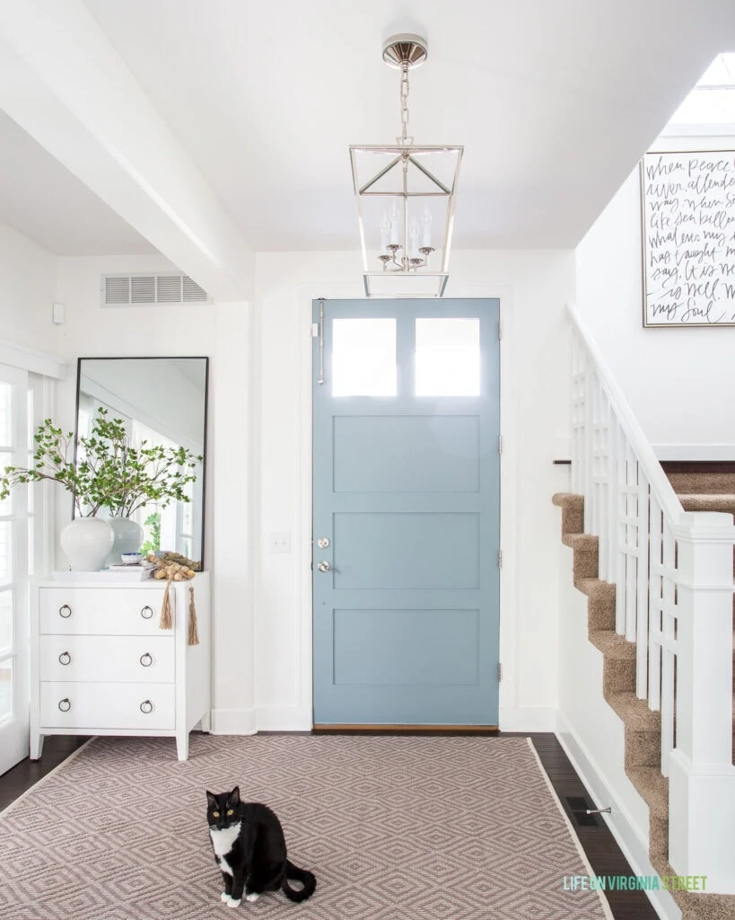A light and bright entryway with a neutral rug in a diamond pattern, white cabinet, tall leaning mirror, blue front door, and a lantern pendant light.