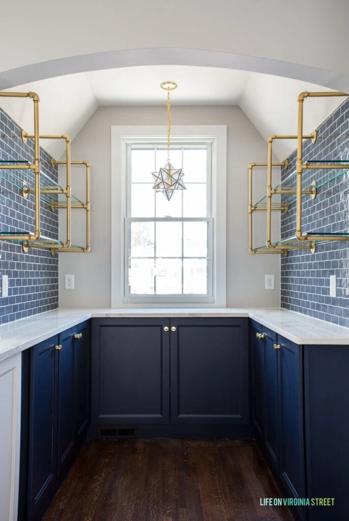 A kitchen butler's pantry with Sherwin Williams Naval cabinets, gold pipe & glass shelves, a Moravian star pendant light, and white quartz countertops.