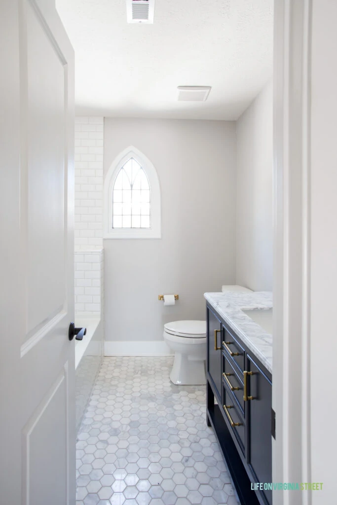 A hallway bathroom with marble hex floors, white beveled subway tile shower, navy blue vanity with Carrara marble top, original leaded pane arched window, and gold accents.