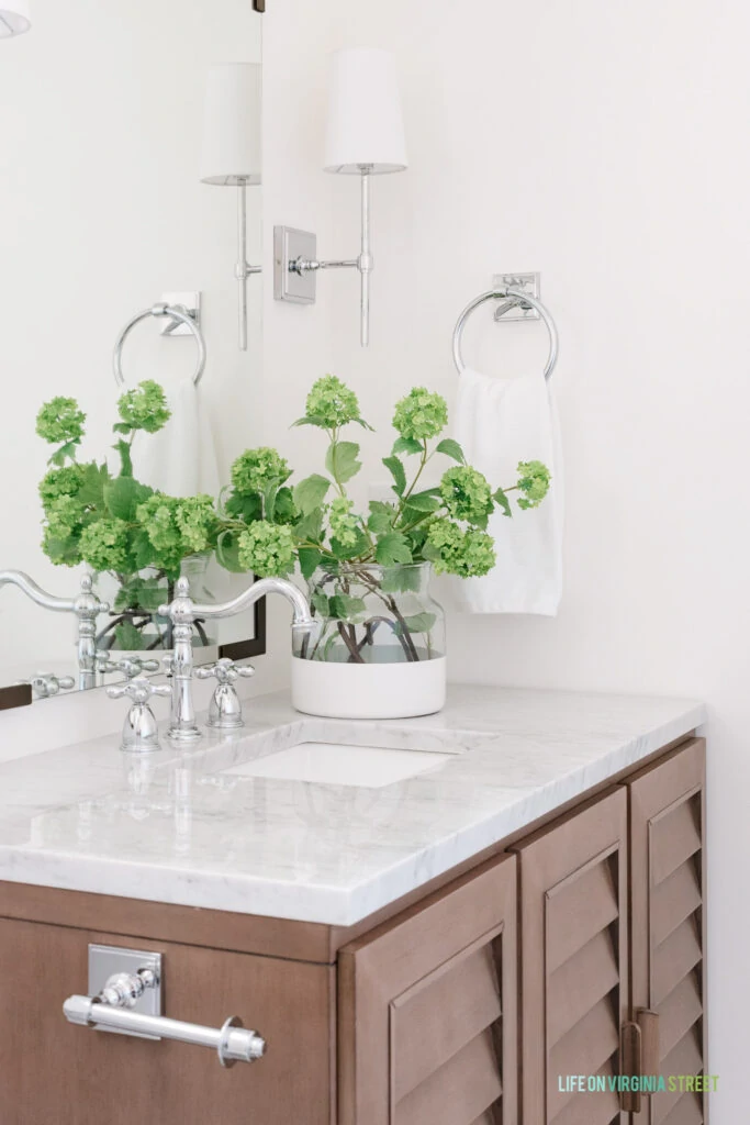 A louvered wood vanity with Carrara marble countertop, chrome toilet paper holder, silver towel ring, vintage style faucet and chrome sconce light fixtures.