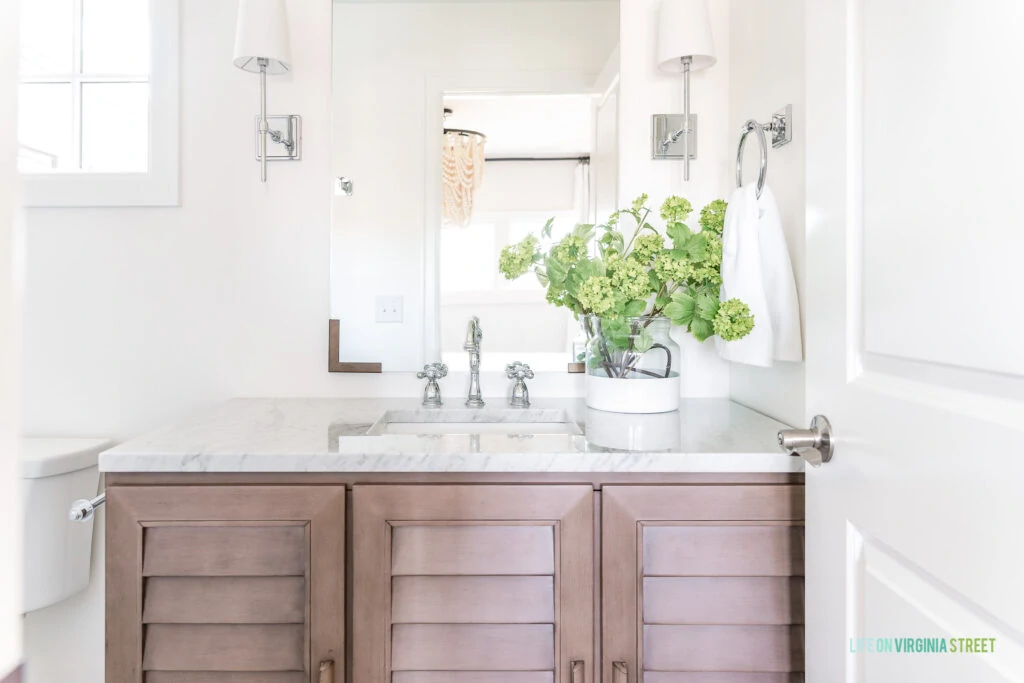 A wood shutter vanity cabinet in a small guest bathroom. Includes a Carrara marble countertop, chrome faucet, chrome sconce lights, and paint dipped vase filled with viburnum stems.