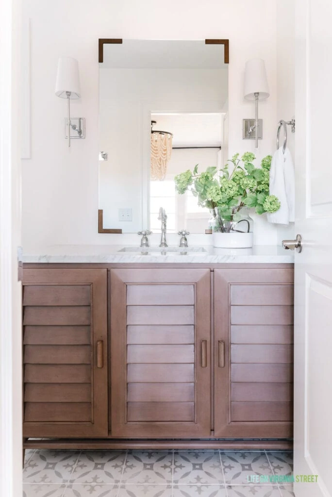 Small guest bathroom remodel with a louvered wood vanity, porcelain tile that looks like patterned cement tile, chrome sconce lights, and a mirror with metal corners.