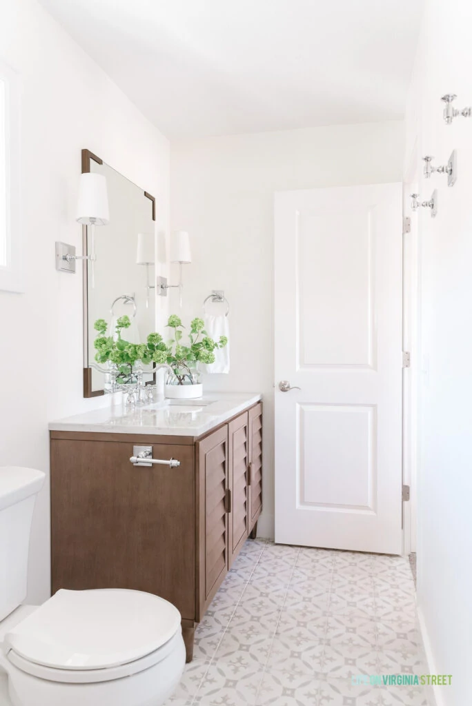 A small guest bathroom remodel featuring a louvered wood vanity, neutral patterned tile floors, chrome sconce lights, a tall vertical mirror with bracket corners, and chrome robe hooks on the wall for towels.