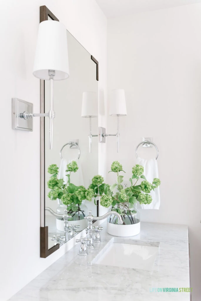 A Carrara marble bathroom countertop with faux viburnum stems, chrome sconce lights and a large vertical mirror.