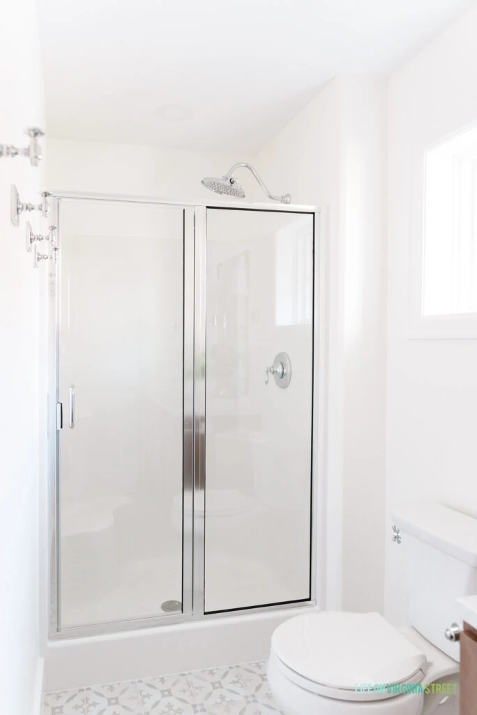 A chrome framed shower door in a shower insert. Also shown is a small square window, cement tile looking floors, robe hooks and a large shower head.