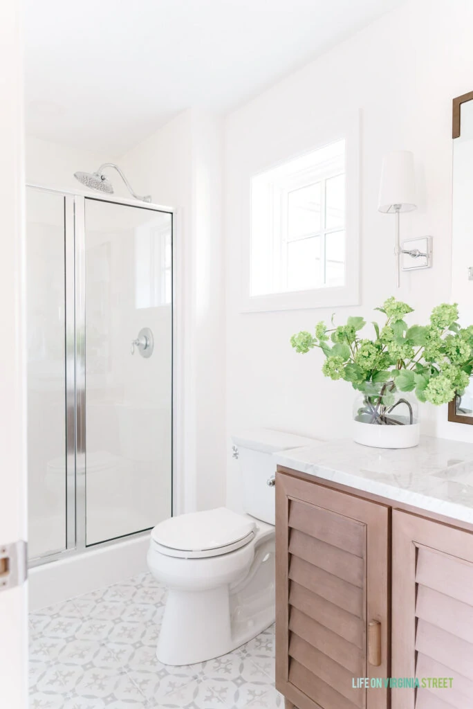 Small guest bathroom remodel with a louvered wood vanity, porcelain tile that looks like patterned cement tile, small square window, silver framed shower door stall, chrome sconce lights, and a mirror with metal corners.