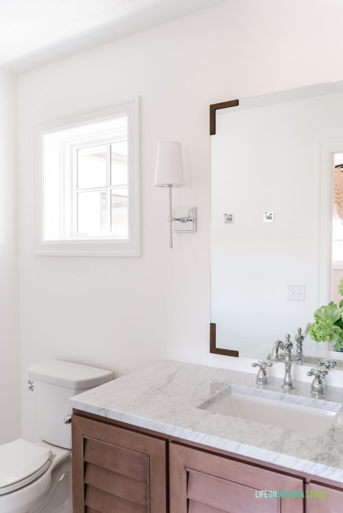 A small bathroom renovation adding a small square window, wood shutter vanity, chrome sconces, vintage style faucets, and a mirror with bronze metal brackets.