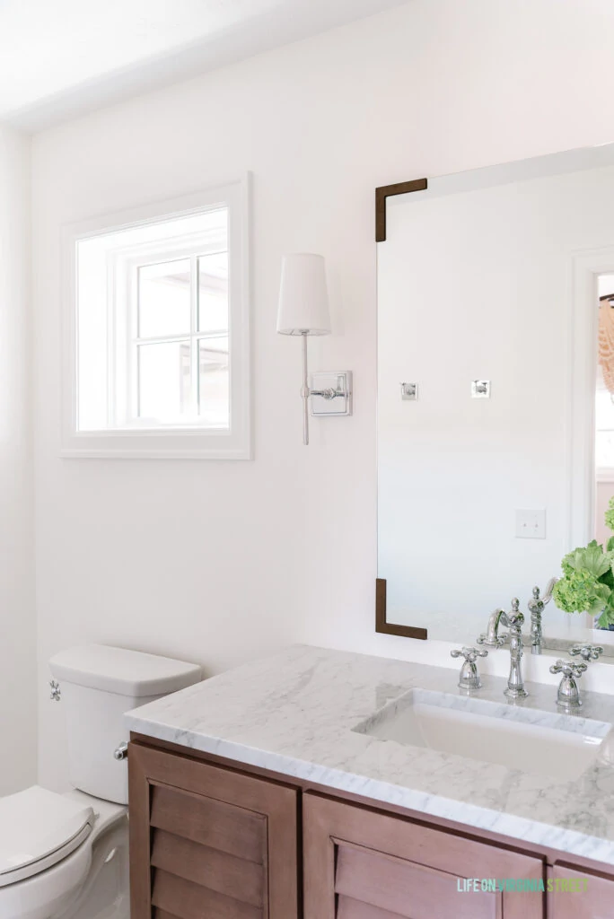 A small bathroom renovation adding a small square window, wood shutter vanity, chrome sconces, vintage style faucets, and a mirror with bronze metal brackets.