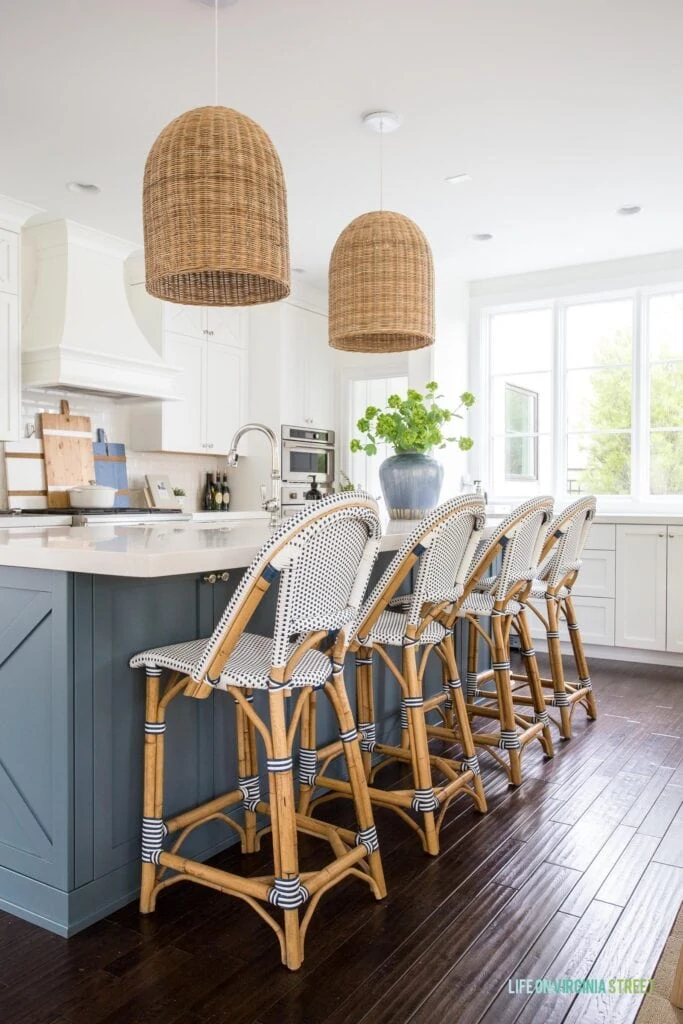 This Serena & Lily Riviera Counter Stool review includes photos of the stool in this coastal style kitchen with white cabinets, a blue island, dark hardwood floors and a large window.