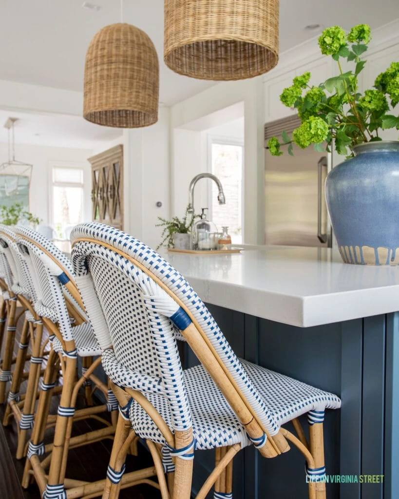 A row of Serena & Lily Riviera Counter Stools in a coastal inspired kitchen with blue island, white quartz countertops, and a large blue ceramic vase filled with faux viburnum.