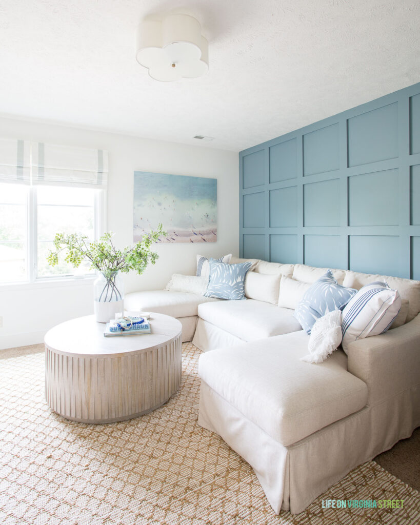 A cozy TV room with blue board and batten grid wall, linen sectional, fluted coffee table, jute rug, beach art, and roman shades.