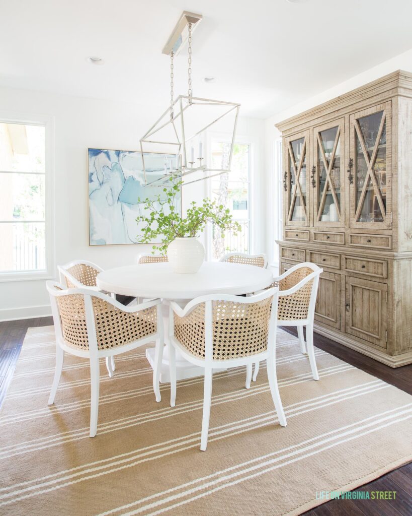 A coastal style dining room with white walls, a white concrete dining table, cane dining chairs, a linear chandelier, wood hutch, striped rug, and blue abstract art.