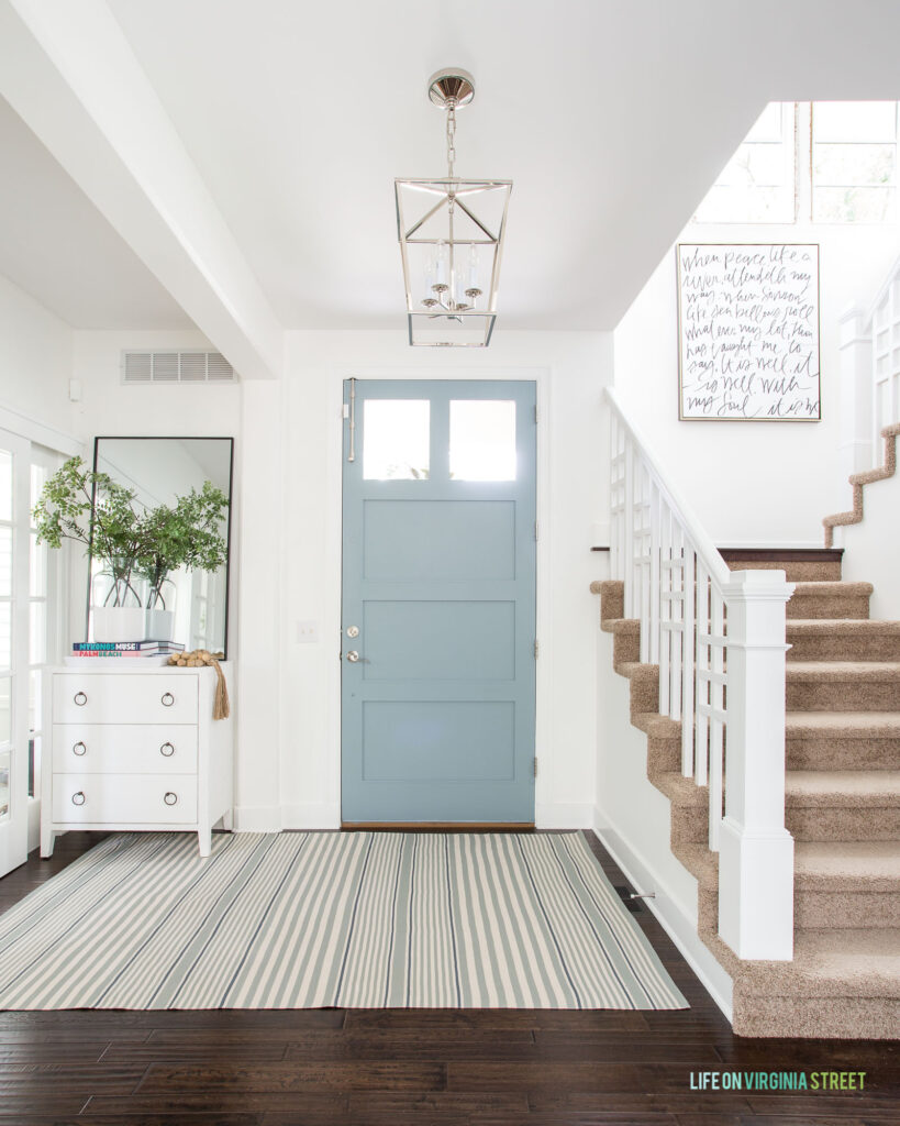 A light and bright entryway with Water's Edge painted front door, striped rug, faux greenery, tall mirror, and lantern pendant light in this 2021 Summer home tour.