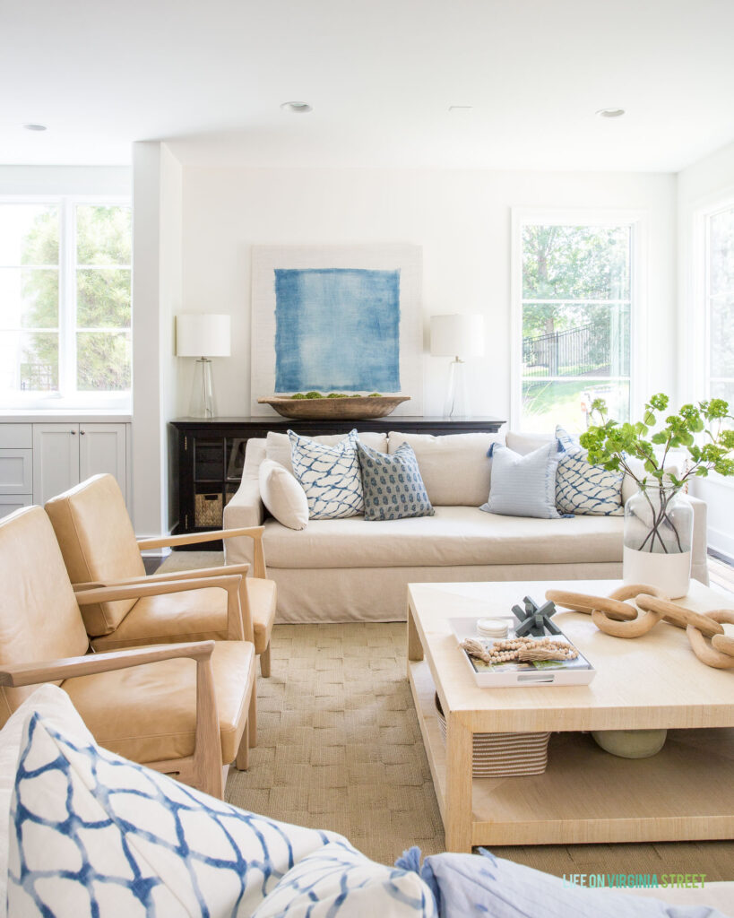 A summer living room featuring an oversized blue and white abstract art piece, leather chairs, linen sofa, raffia coffee table and blue and white pillows in a 2021 summer home tour.