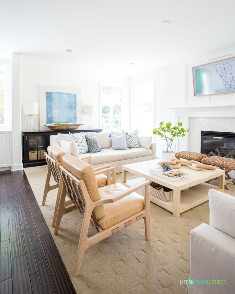 A coastal style living room with a traditional coastal coffee table wrapped in raffia, linen sofas, blue and white pillows, a black cabinet, woven stool, and light leather chairs.