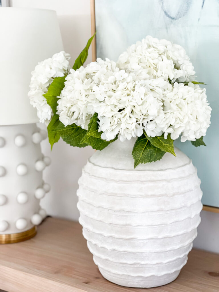 An oversized ridged white ceramic planter filled with faux hydrangeas and decorated in a coastal dining room.