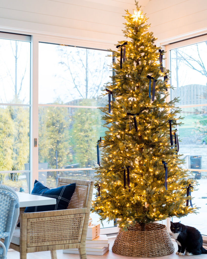 Best Christmas tree collar ideas! An affordable woven tree collar at the base of a 9' artificial Christmas tree decorated with navy blue velvet bows. Also includes a black cat and rattan dining chair.