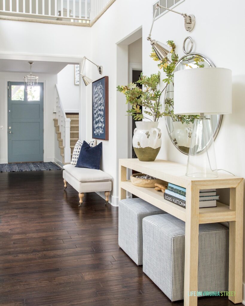 An entryway hallway decorated for fall with dark hardwood floors, a light raffia console table, pillowtop bench, glass lamp and faux oak leaves. 