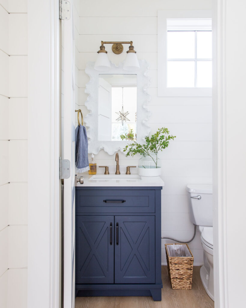 A small pool house bathroom with white shiplap walls, navy blue vanity, white coral style mirror, gold light fixtures and faucet, and a square window.