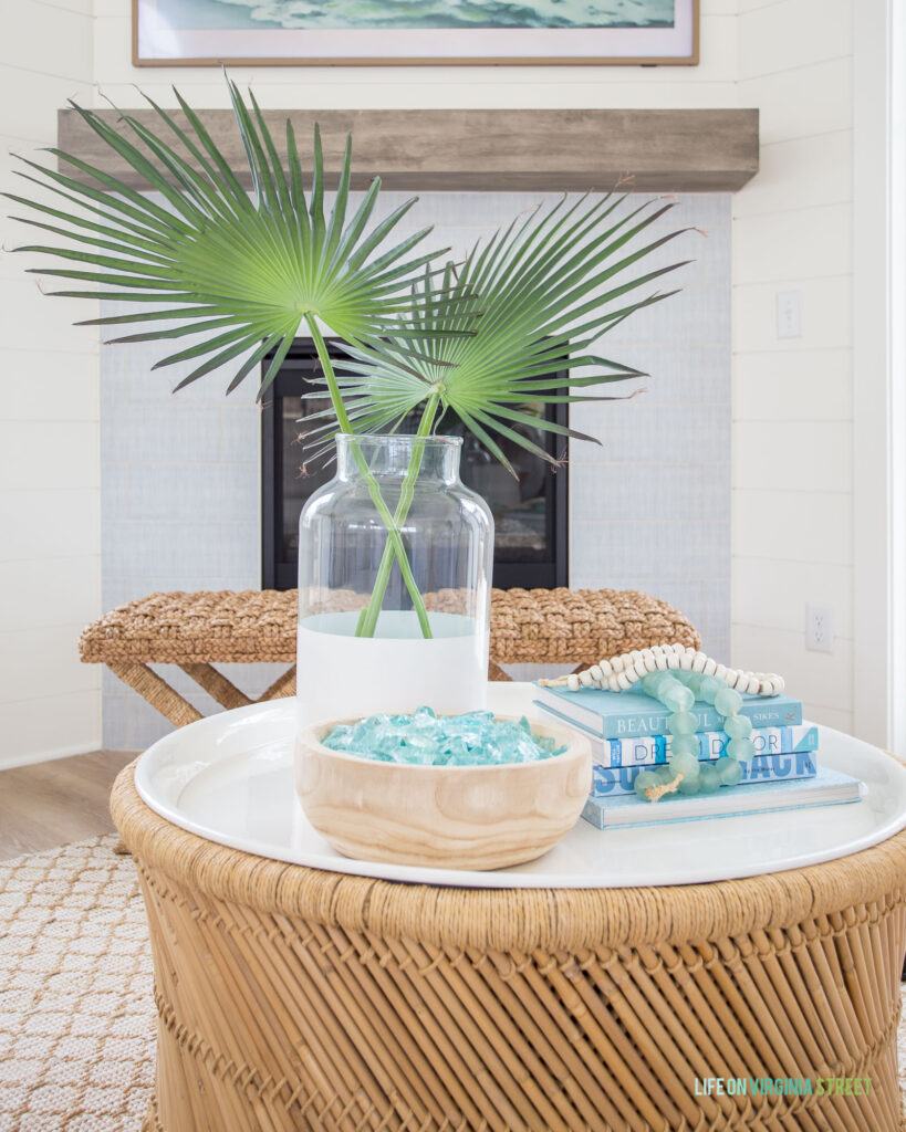 A round bamboo coffee table with white enamel top and decorated with sea glass, palm leaves, recycled glass beads and coastal coffee table books.