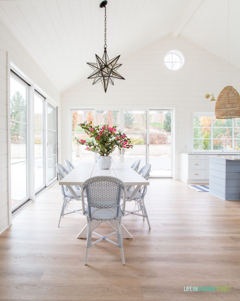 A pool house dining area with a long white picnic table, bistro chairs, light LVP floors, a large Moravian star light, white shiplap walls, and large sliding glass doors.