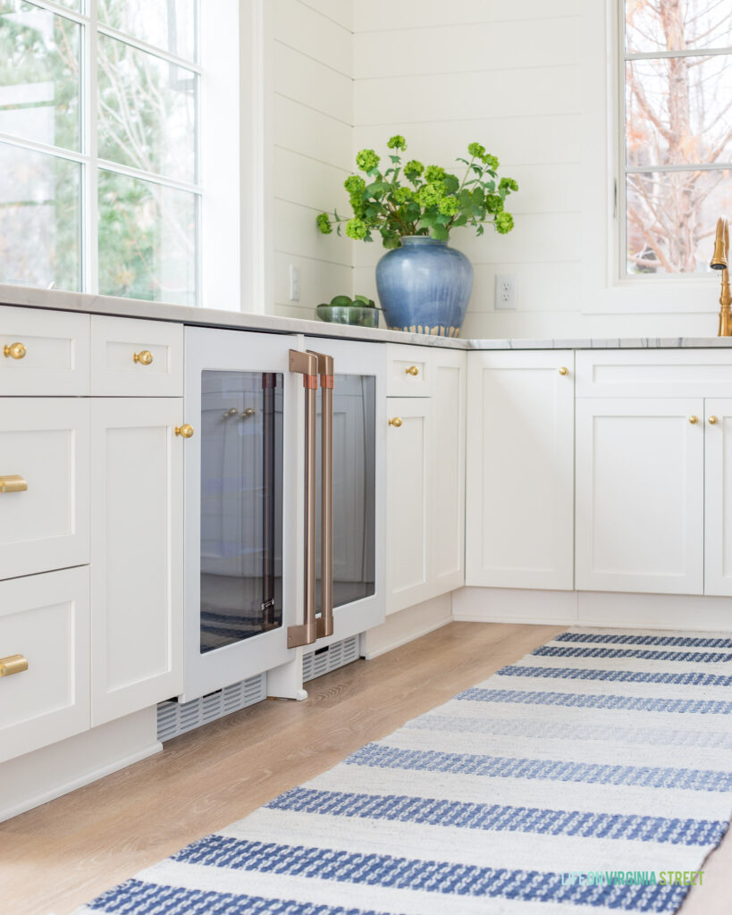 Cafe Appliances Beverage Fridges in a pool house kitchen with white cabinets, gold hardware, a blue and white striped rug, and white shiplap walls.