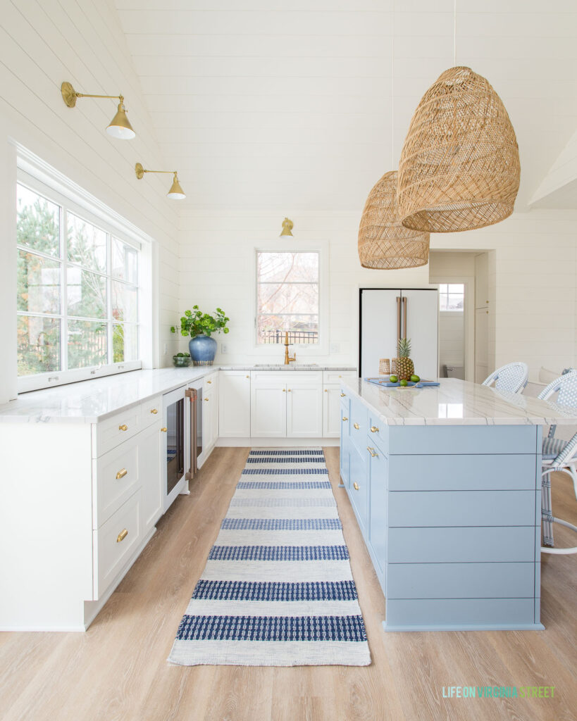 A pool house kitchen decorated with white perimeter cabinets, a blue island, blue and white striped rug, basket pendant lights, and white shiplap walls.