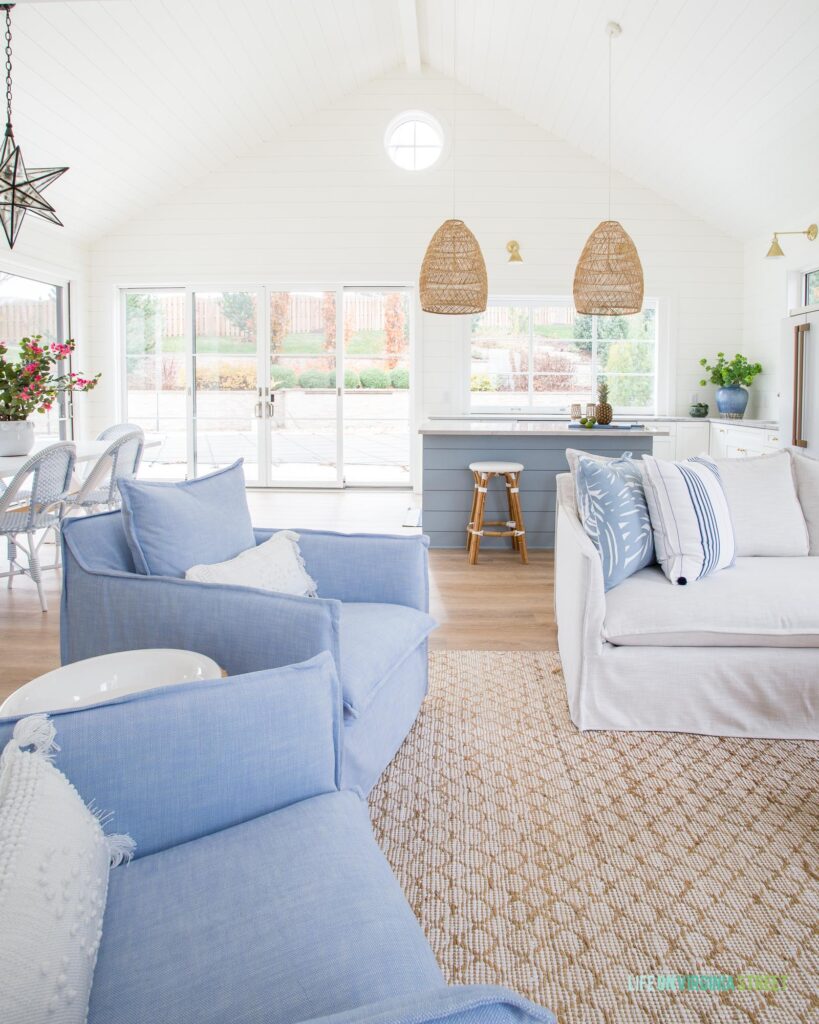 An enclosed pool house with white shiplap walls, vaulted ceiling, round circle window, blue and white furniture, a blue kitchen island and rattan basket pendant lights.