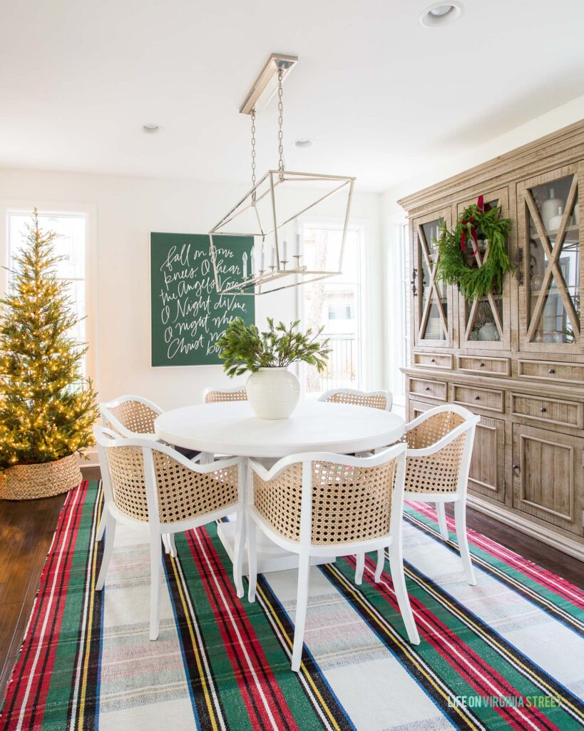 A woven seagrass tree collar in a Christmas dining room with a round white table, cane chairs, a Stewart plaid rug, a wreath tied to the hutch, and oversized Christmas art.