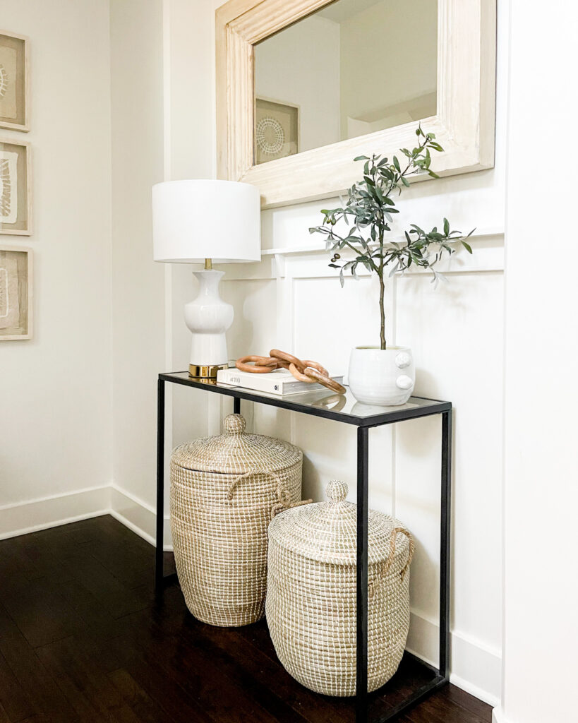 A mini entryway makeover with large woven baskets, a linen covered coffee table book, a white ceramic lamp, wood chain decor, black console table and large whitewashed mirror.