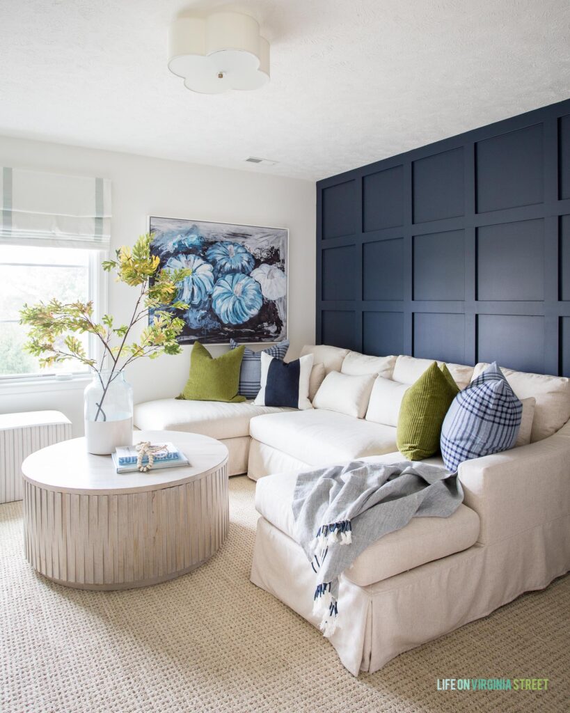 A Pottery Barn linen sectional and round wood coffee table in a den with a navy blue board and batten grid wall.