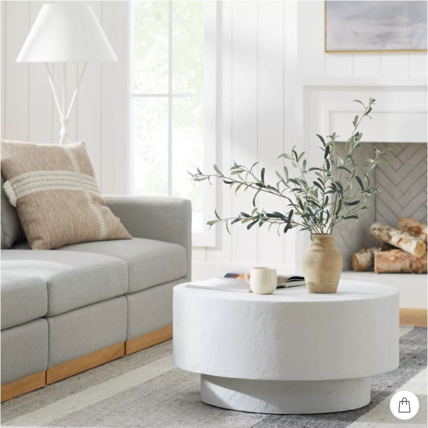 Simply living room decor with a gray modular sectional, round white concrete style coffee table, white tripod lamp, and faux olive stems.