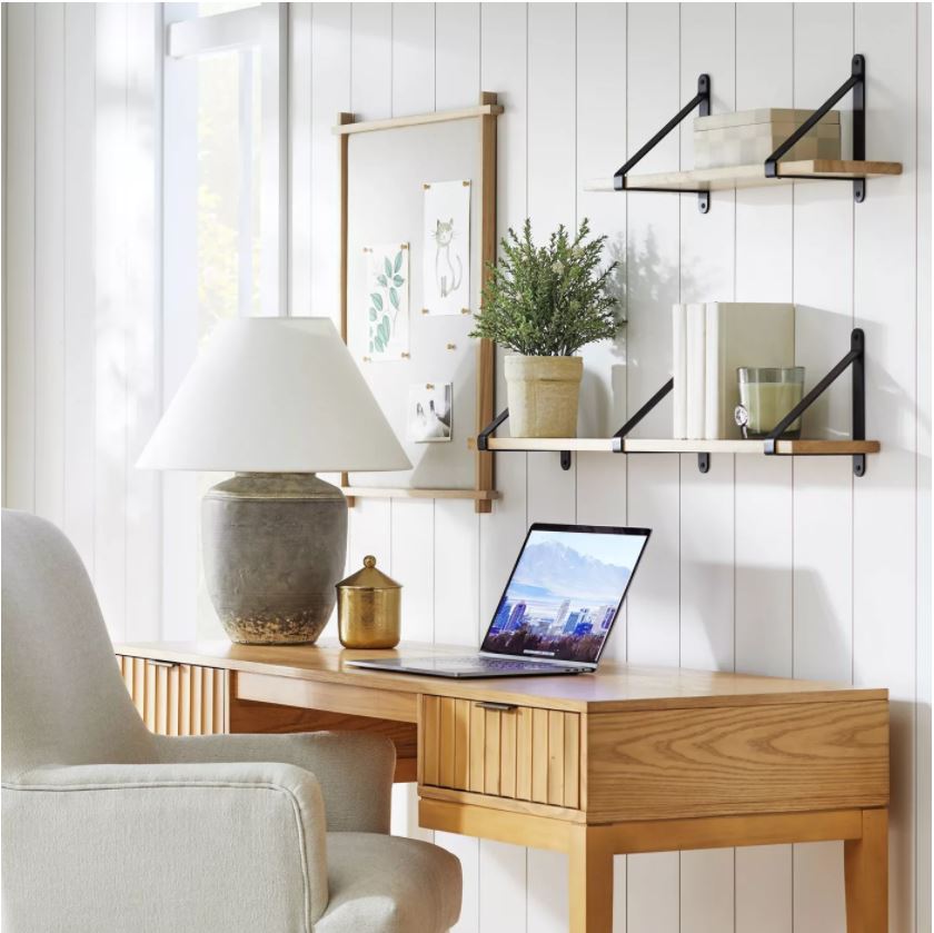 A small home office desk space with a scalloped desk, upholstered desk chair, hanging shelves, a linen pinboard, and dark ceramic table lamp.