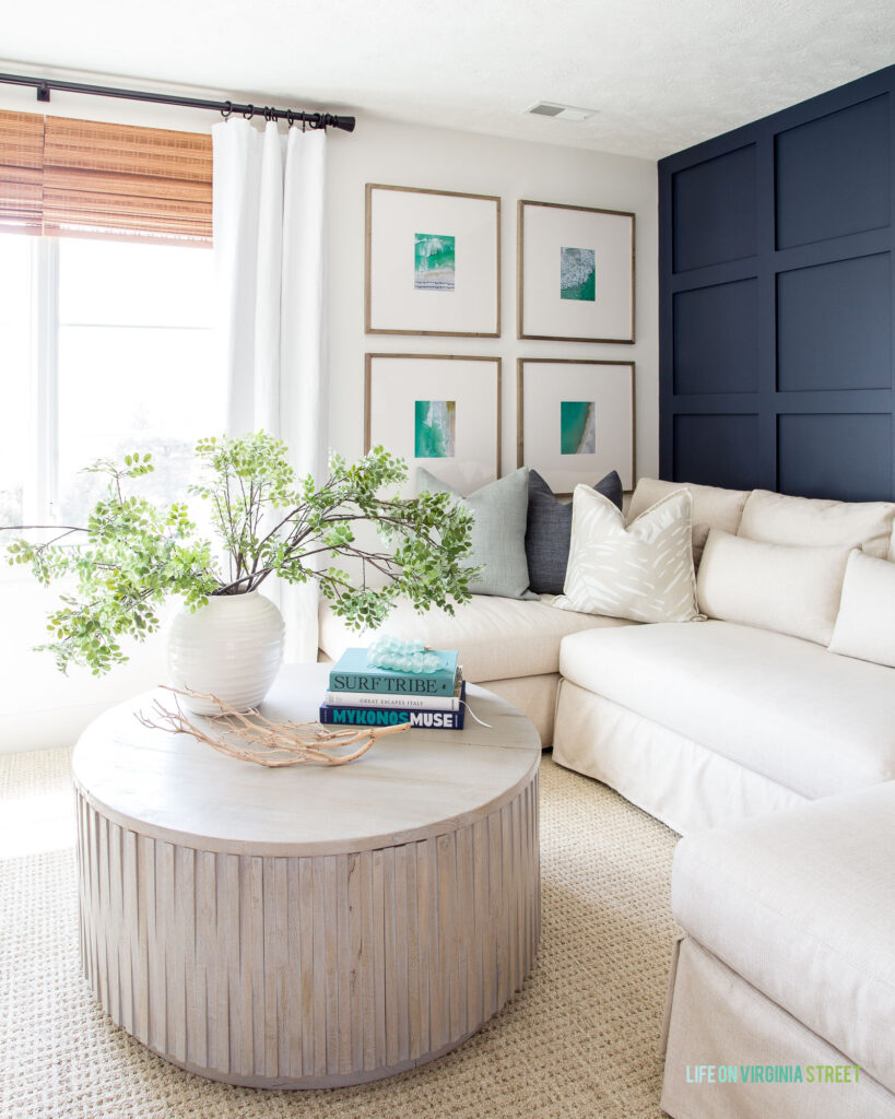 A cozy living room scene with a linen sectional, round wood coffee table, beach art gallery wall, and a navy blue board and batten accent wall.