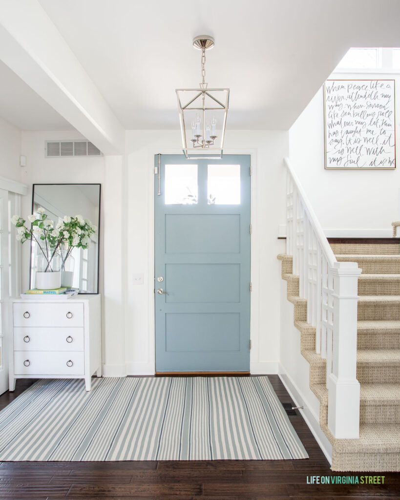 The Life On Virginia Street 2022 Spring Home Tour, including the entryway with a light blue front door, striped rug, white raffia cabinet, and a staircase leading up to word art.