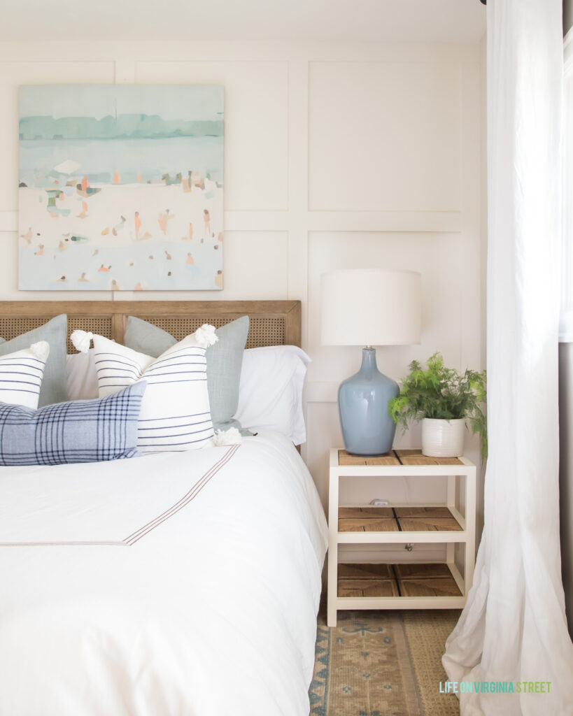 A guest bedroom with beach art hung above a cane bed with white bedding, striped pillows, a blue ceramic lamp, and hand-knotted rug.
